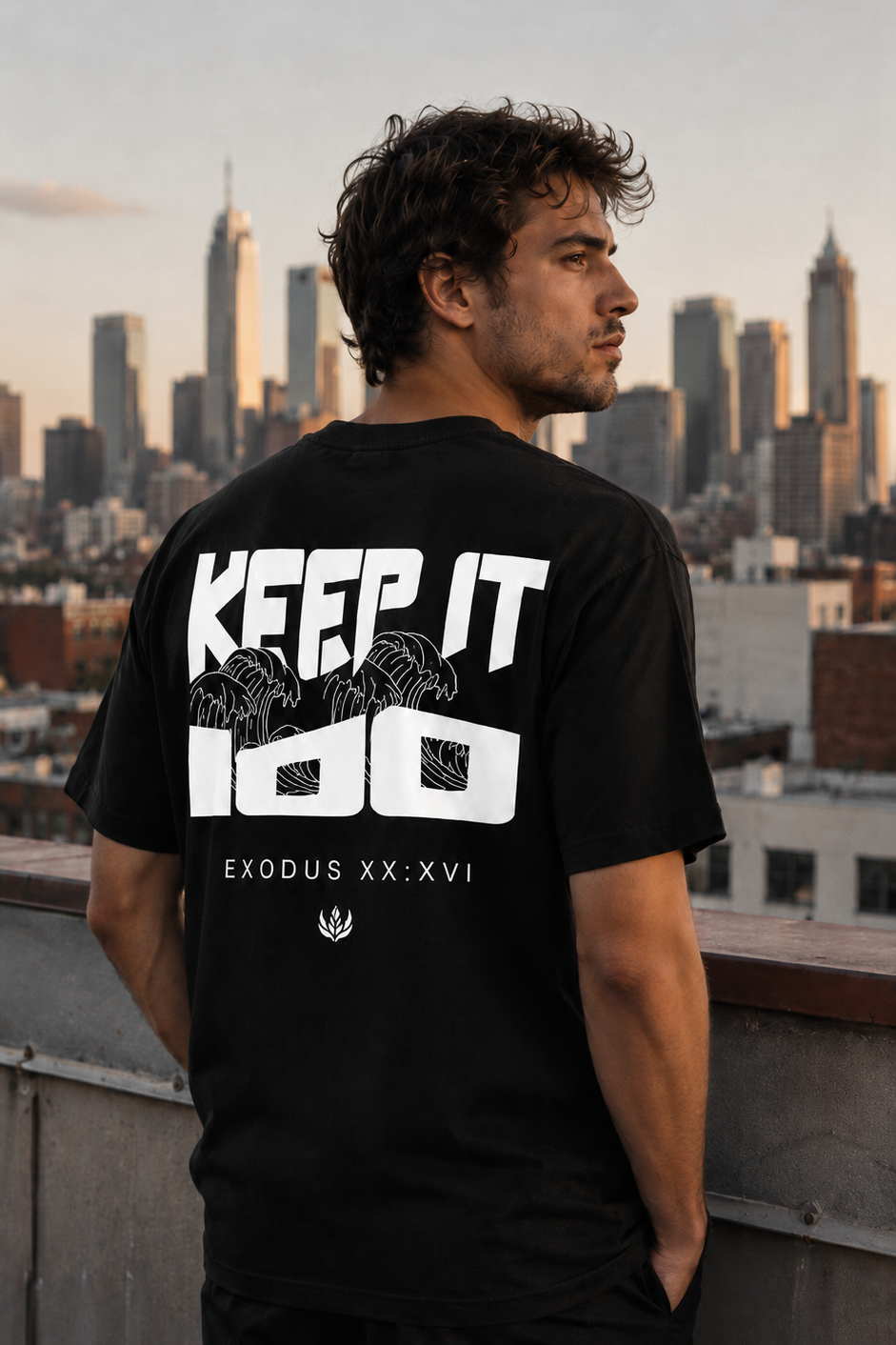 Man wearing a black t-shirt with 'KEEP IT 100' text, standing on a rooftop with a city skyline in the background.